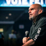 Fetterman Shocks Dems–Shakes Trump Hand Man speaking into microphone on stage.