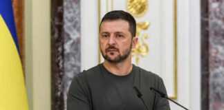 Man speaking at podium with microphones, Ukrainian flag behind.