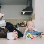 A tired parent with their head down on the table next to a curious baby