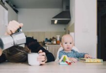 A tired parent with their head down on the table next to a curious baby