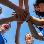 Group of female athletes joining hands in a circle under a blue sky