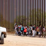 Unvetted Sponsors: Migrant Kids in Grave Danger Group of children and adults walking near a border wall with a customs vehicle