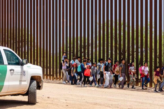 Group of children and adults walking near a border wall with a customs vehicle