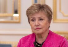 A woman in a pink jacket with a serious expression, seated in a formal interior