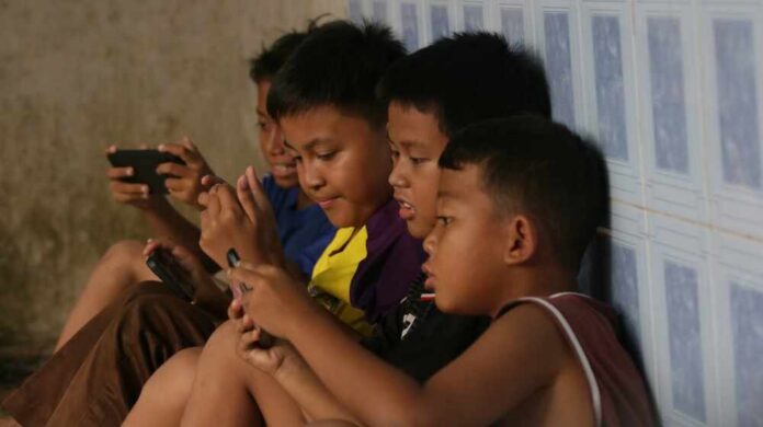 Group of children sitting against a wall, focused on their smartphones