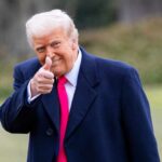 A man in a dark coat and pink tie giving a thumbs up outdoors
