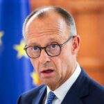 A man in a suit with glasses, looking serious in front of an EU flag