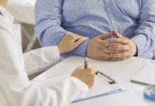 A doctor writing notes while a patient sits with hands clasped, receiving support.