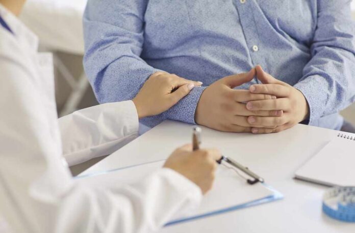 A doctor writing notes while a patient sits with hands clasped, receiving support.