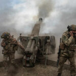 Soldiers fire a field howitzer as smoke and dust rise around them