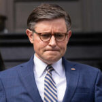 Man in blue suit with glasses looks down thoughtfully