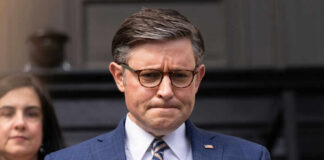 Man in blue suit with glasses looks down thoughtfully