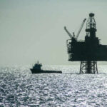 An offshore oil rig silhouetted against the shimmering sea with a small boat nearby