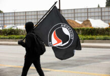 Person in black hoodie carrying a large black flag while walking on a street