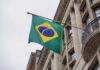 Emergency System Compromised — Brazil In The Dark A hand holding a smartphone displaying a severe weather alert against a stormy background