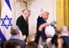Two suited men at a formal event with an Israeli flag and audience