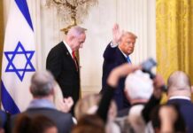 Two suited men at a formal event with an Israeli flag and audience