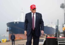 A man in a suit wearing a red cap stands near a ship at a harbor