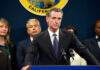 Newsom World Rocked By Co-Conspirator Person speaks at podium with California seal behind.