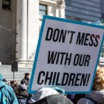 Strict Liability Tsunami Targets Gender Clinics protest sign held in a crowd outside a government building