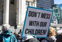 Strict Liability Tsunami Targets Gender Clinics protest sign held in a crowd outside a government building