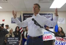 man in white shirt speaking at a rally with supporters and signs behind him