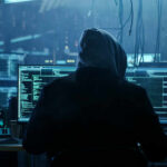 Teen Hacker Drains $100M — Extradited A person in a hoodie working on multiple computer screens in a dimly lit room