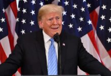 Ballot Fight Erupts: Trump Votes By Mail man at podium with U.S. flags behind him