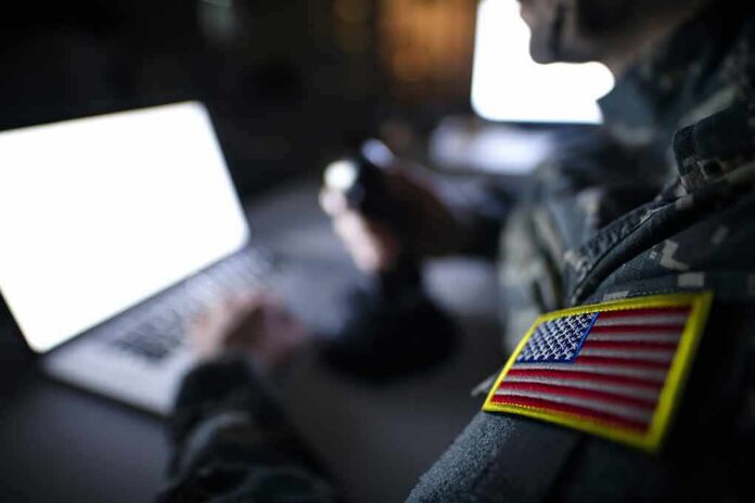 shutterstock_1395036650 Soldier with U.S. flag patch using a laptop in a dim room