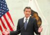 Newsom’s Power Play Hits Fresno man in suit speaking at podium between U.S. and state flags