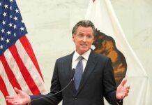 Newsom’s Power Play Hits Fresno man in suit speaking at podium between U.S. and state flags