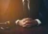 person in suit with hands clasped at a desk beside a gavel