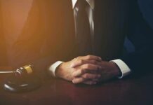 person in suit with hands clasped at a desk beside a gavel