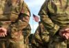 Soldiers in camouflage stand with hands behind backs facing a U.S. flag
