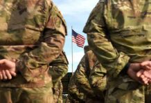 Soldiers in camouflage stand with hands behind backs facing a U.S. flag