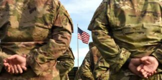 Soldiers in camouflage stand with hands behind backs facing a U.S. flag