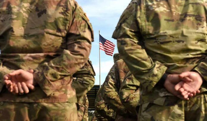 Soldiers in camouflage stand with hands behind backs facing a U.S. flag