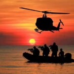 Cold, Pain, Laughter – SEAL Survival Code Helicopter hovers above armed team in inflatable boat at sunset