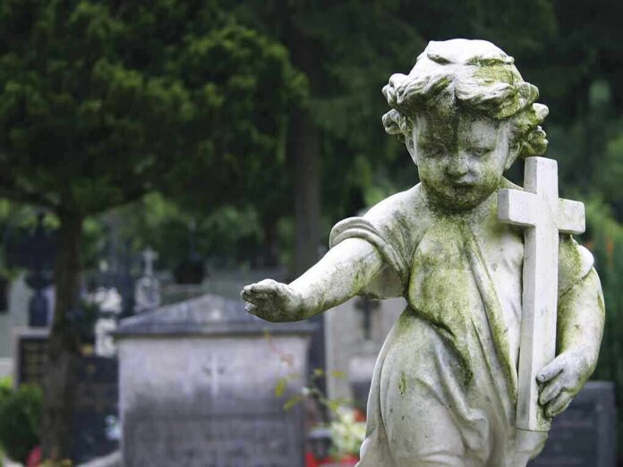 shutterstock_156593303 Weathered statue of a child angel holding a cross in a cemetery